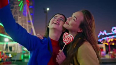 Gülen kızlar aydınlatılmış lunaparkta selfie çekiyorlar. Lunapark gecesinde cep telefonuyla fotoğraf çeken iki arkadaş. Mutlu güzel kızlar, neon atlıkarıncada iyi eğlenceler.