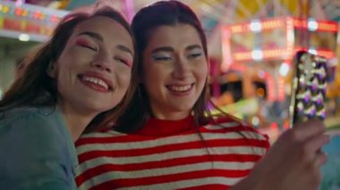 Kızlar lunaparkta komik selfie çekiyorlar. Neşeli en iyi arkadaşlar renkli lunaparkta eğleniyorlar. Mutlu gülümseyen kız kardeşler akıllı telefon fotoğrafları çekiyor. Arkadaşlık bağı kavramı