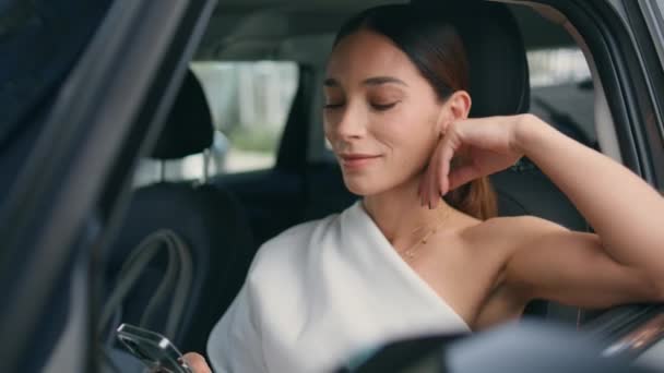 自信的女士坐在汽车前排 微笑着看着智能手机屏幕 成功的女商人在昂贵的汽车里给手机留言 年轻的女司机在停车场休息 — 图库视频影像