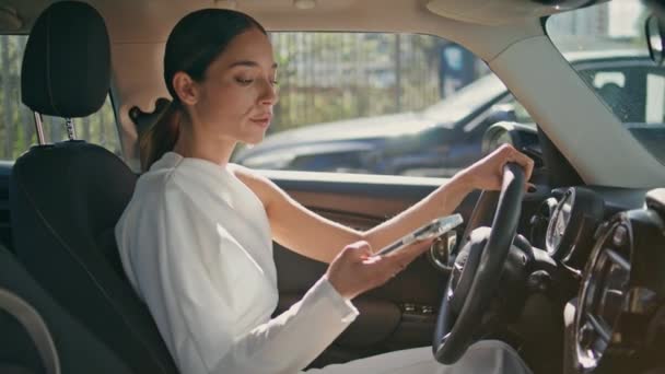 高価な車に乗っている自信のある女性ドライバーは スマートフォンのメッセージを閉じています シックな若いビジネスマンオープン自動車ミラー 市の駐車場に立っているVip自動車の中にポーズする魅力的な少女 — ストック動画