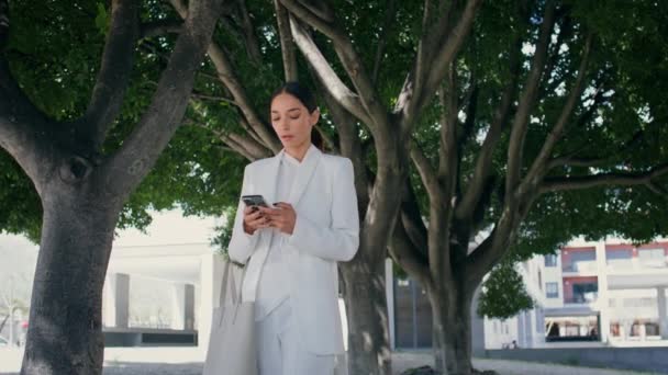 女商人在美丽的绿树下走着 拨电话号码 穿着白色西服的优雅时髦的女商人在户外打电话 非常漂亮的女人用智能手机进行工作交流 — 图库视频影像