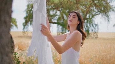 Çayıra çamaşır asan şehvetli kadın. Romantik esmer kadın temiz beyaz çarşafları ipe geçirip çavdar tarlasında çamaşır yıkamanın keyfini çıkarıyor. Fotoğrafçı kız kırsalda poz verirken kameraya bakıyor.