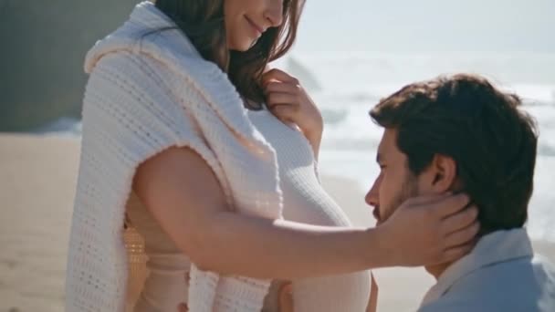 丈夫抱着怀孕的肚子蹲在幸福的妻子身边 站在美丽的沙滩特写上 充满爱心的女人带着爱心看着未来的父亲 温柔体贴的新婚夫妇在海滨生孩子 — 图库视频影像