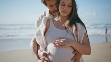 Sevecen hamile çift güzel güneşli sahilde poz veriyor. Yakın çekim. Sevgi dolu koca, rahat karısının arkasında durup koca göbeğine sarılıyor. Romantik sakin eşler hafta sonları deniz kıyısında dinleniyorlar..