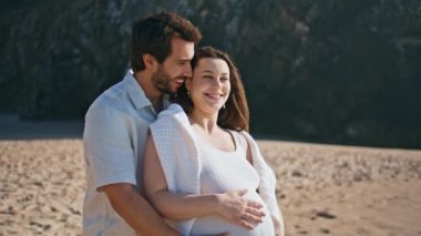 Gülümseyen adam hamile göbeğini kucaklıyor sevgili genç karısı kumlu deniz kıyısında güzel bir şekilde dikiliyor. Mutlu kaygısız evli çift yaz plajında rahatlarken hamileliğin tadını çıkar. Neşeli aile neşe içinde.