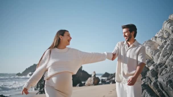 妊娠中の配偶者が日当たりの良い岩のビーチを歩く笑い 喜びを感じる幸せが近づいています 夏の海岸で一緒に楽しんでいる恋愛の幸せなカップル 陽気なひげ 愛する — ストック動画