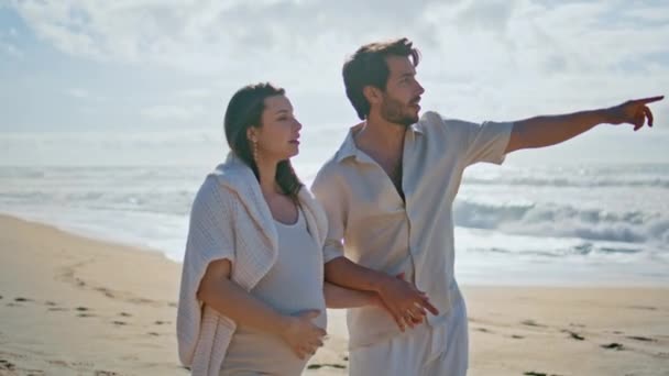 妊娠中のカップルは ゴールデンビーチの砂の上を歩く海の風景を楽しんでいます 愛する妻に自然の美しさを示す幸せな柔らかい夫 海岸散歩でハンサムな男を抱擁する赤ちゃんを期待するケアフリーの女性 — ストック動画