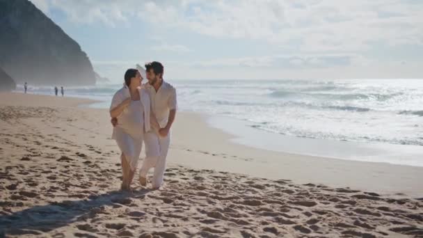未来の両親は 一緒に笑う美しいビーチを歩いています 晴れた海岸でロマンチックな夜を過ごす若いカップルを愛する 泡立った海の波の近くを散策するロマンチックな妊婦の家族 — ストック動画