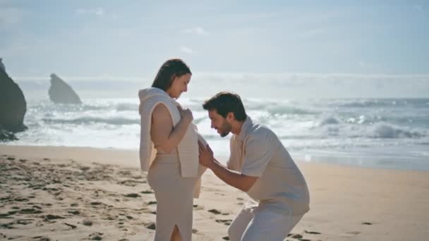 妊娠中のカップルは美しい海の波の正面に立っている日 愛する夫は優しさと大きな女性の腹にキスします 海岸でロマンチックな週末を過ごす赤ちゃんを待っている幸せな結婚した配偶者 — ストック動画
