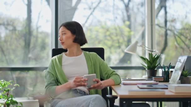 オフィスの閉鎖でコーヒーを飲むアジアの女性 落ち着いたスタートスーパーガールは コンピュータの職場で休む距離を見ています パノラマウィンドウでクリエイティブなコワーキングに座っているドリーミーな女性 — ストック動画