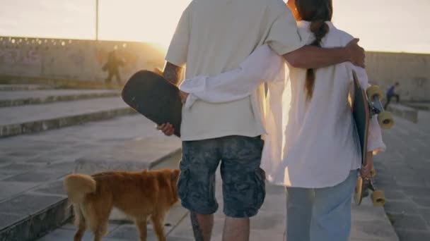 爱滑冰的人在阳光下漫步 在体育场后视镜 两个年轻的恋人遛狗穿过夏天的街道 男友情侣拥抱在阳光下走到户外 — 图库视频影像