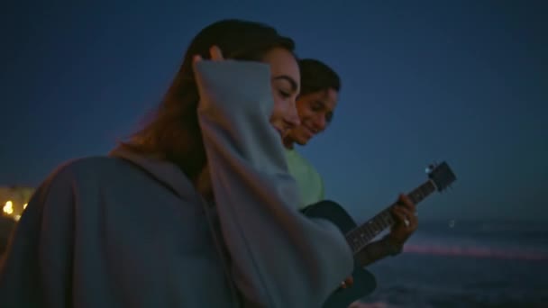 与夜间海滩特写约会的忧郁伴侣 放松的无忧无虑的女人喜欢音乐 年轻的男人晚上在海边弹吉他 两名青少年在暮色的夏日里尽情放松 — 图库视频影像