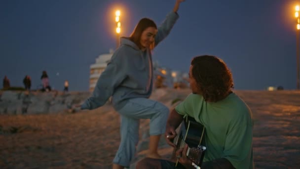 晚上沙滩上拥抱男朋友的年轻女人 一个英俊的男人在暮色中弹奏吉他 浪漫的情人在度假时一起休息 听着音乐的快乐女士 — 图库视频影像