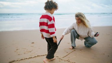 Anne oğul sonbahar haftasonu sopalarla kumsal çiziyor. Tatilde kumsal yazan bir aile. Güzel anne bebek bakıcısı sevimli kıvırcık çocuğa öğretiyor. Ebeveynlik yaşam tarzı çocuk bakımı kavramı.