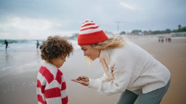 Anne çocuk sonbahar haftasonunda deniz kenarında oynuyor. Okyanus manzarasında taş tutan güzel bir kadın tatlı kıvırcık çocukla konuşuyor. Güzel bir aile birlikte yaratıcı oyun oynuyorlar. Bakımlı yetiştirme konsepti.