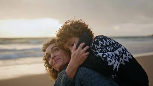 Cute kid talking phone on coast vacation with mother closeup. Happy family speak smartphone enjoying distance communication with father weekend travel. Curly son hug mom resting sunset ocean together.