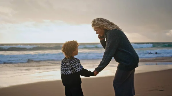 Mother kid resting shore on autumn sunset together. Smiling woman talking phone calling husband. Adorable curly kid enjoying family weekend having fun on beach. Busy parent businesswoman work holiday