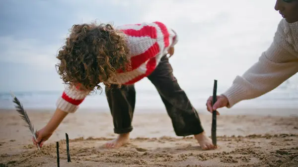 Mom kid drawing beach sand at cloudy sky. Creative family having fun together resting seaside weekend. Beautiful curly mother babysitter teaching helping adorable boy. Parenting learning concept.