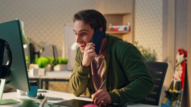 Ofiste telefonla konuşan neşeli bir adam. Kapatın. Yaratıcı iş yerindeki akıllı telefondan ilham alarak çalışan mutlu tasarımcı. Gülümseyen yaratıcı dolapta dijital kalem kullanarak tablet çiziyor