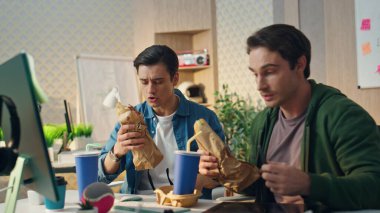 İş yerindeki iş adamları iş yerinde konuşurken sandviç yiyorlar. Duygusal adamlar bilgisayar karşısında öğle yemeği molasında yemeğin tadını çıkarıyorlar. Modern hipster ofisinde iletişim kuran iki ortak.