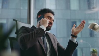 Başarılı yatırımcı akıllı telefondan konuşuyor. Lüks ofis kapanıyor. Mutlu sakallı girişimci telefon konuşmasında gülerek iyi haberler alıyor. İş yerinden rahatlamış işadamı çağrısı.