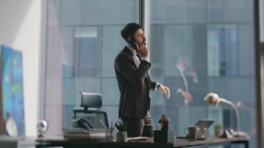 Zengin işyeri sahibi panoramik pencereye yaslanarak lüks ofisi arıyor. Kendine güvenen sakallı iş adamı, modern işyerindeki akıllı telefondan iş görüşmesi yapıyor. Başarılı bir yönetici telefonda konuşuyor