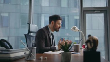 Ofis masasındaki dizüstü bilgisayarla çevrimiçi toplantıda kendine güvenen bir yönetici danışmanlık yapıyor. Modern işyerinde oturan müşteriye pozitif motivasyonlu danışman videosu. Mutlu sakallı iş adamı sohbette konuşuyor..