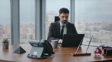 Laptop 'ta çalışan kendine güvenen iş adamı lüks ofisinde oturuyor. Odaklanmış sakallı adam, ahşap masada modern bilgisayarda yazı yazıyor. Ciddi zengin şirket sahibi iş stratejisi oluşturmakla meşgul..