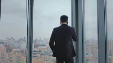 Şirket yöneticisi panoramik ofis penceresinden ortakları arıyor. Kendine güveni tam bir iş adamı akıllı telefonda iş meselelerini çözüyor. Zarif takım elbiseli arka plan çalışanı içeride telefonla konuşuyor..