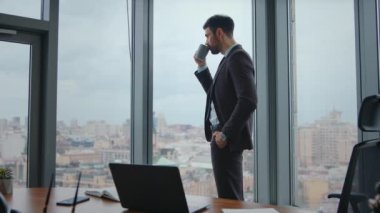 Kendine güvenen yönetmen panoramik ofis penceresinde içki içiyor. Ciddi sakallı yönetici elinde fincanla lüks işyerinde kahve molası veriyor. Şehir manzarasına bakan girişimci..