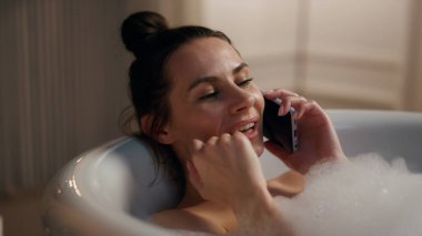 Romantik manken akıllı telefon soğutma küvetini kapatıyor. Sıcak kabarcıklar döşeyen rahat mutlu bir kız banyoda flört eden bir cep telefonuyla. Gülümseyen kadın evde spa prosedürlerinden zevk alıyor. Evcil güzellik rutini