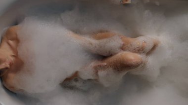 Genç bayan köpüklü küvet manzarasını rahatlatıyor. Rahatlamış bir kadın akşam evde tek başına banyo yapıyor. Sakin manken çıplak bacaklara dokunuyor ve rahat bir yerde dinleniyor. İç kaplıca ve refah konsepti