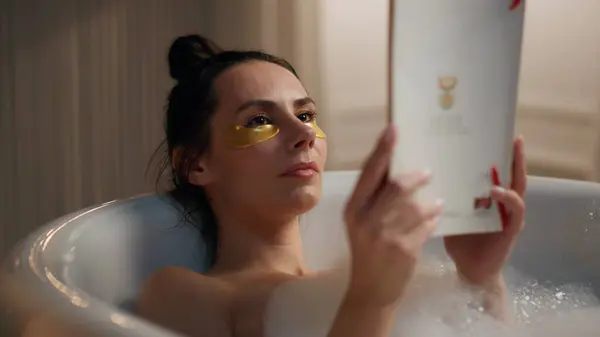 Chilling lady read novel enjoying spa procedures at bath. Closeup calm woman laying bathroom holding storybook at night home. Under eye patches model involved in hobby at hotel. Dreamy girl relaxing