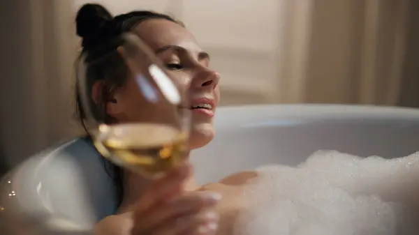 Carefree girl testing alcohol resting bathtub at evening. Lazy woman drinking champagne holding wineglass at hotel. Sensual model taking bath enjoying spa. Luxury lifestyle and hygiene activities