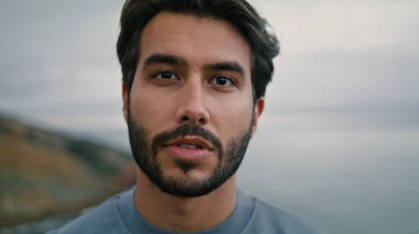 Bulutlu deniz manzaralı arka planda kameraya bakan çekici sakallı adam. Bulutlu deniz kıyısında tek başına duran yakışıklı gencin portresi. Sakin, koyu renk saçlı, kasvetli bir adam..