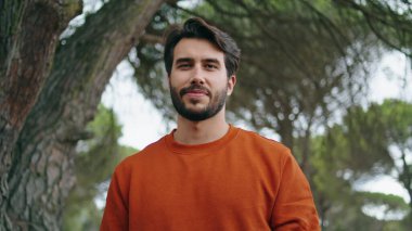 Yeşil Park 'ta kameraya gülümseyen yakışıklı sakallı adam. Sırt çantası taşıyan çekici bir gencin portresi doğada gezintiye çıkıyor. Güzel orman ağaçlarının önünde tek başına duran mutlu turist..