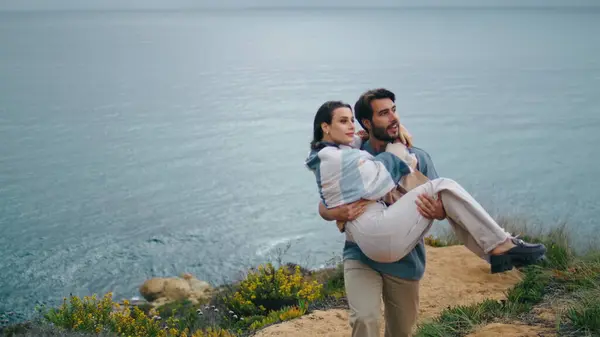 Happy bearded man holding relaxed woman on hands walking at gloomy sea shore. Romantic couple going at coast hill in front gray calm ocean. Loving boyfriend carrying girlfriend on romantic weekend.