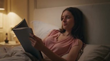 Geceleri sakin bir kadın kitap tutuyor. Hafta sonları evde kitap okuyan gülümseyen kız. Huzurlu genç bayan okuyucu rahat yatakta sakin bir akşamın tadını çıkarıyor. Uyum hobisi gevşeme konsepti.