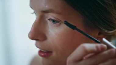 Maskara tutan, kirpiklerini fırçalayan yakın plan kadın. Gülümseyen kadın evde yüzüne hayran. Güzel, rahat bir kadın sabah işe gitmek için günlük makyajını yapıyor. Kozmetik güzellik rutin konsepti.