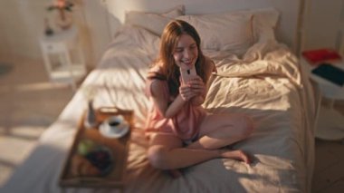 Güneşli yatak odasında akıllı telefon tutan mutlu bir genç. Rüya gibi model, rahat bir yatakta cep telefonunu kontrol ederek sabahları eğleniyor. Romantik gülümseyen kız pijamalarını giyer. Tasasız eğlence konsepti