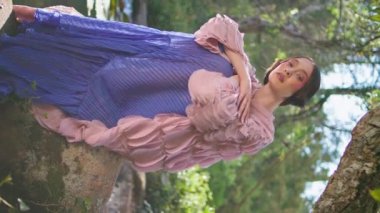 Sihirli kadın orman göletinin yanında oturarak doğal sakinliğin tadını çıkarıyor. Peri masallarındaki yeşil ağacın altında poz veren romantik çekici kız. Büyüleyici model peri berrak göl suyu dikey videosunda dinleniyor..