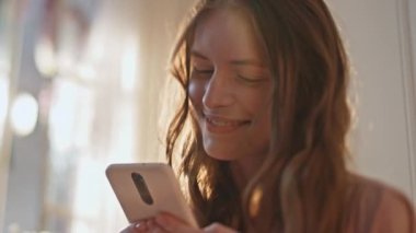 Neşeli kız güneş ışığında mesaj okuyor. Yakın plan. Mutlu genç cep telefonu kullanıyor sosyal medyayı kontrol ediyor ve çevrimiçi video izliyor. Güzel bir kadın arkadaşına mesaj atıyor. Altın sabahta akıllı telefona bakıyor..
