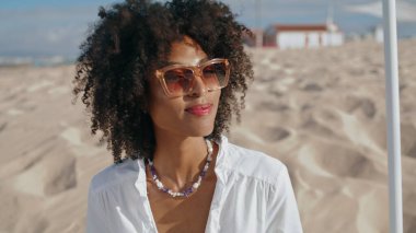 Stylish woman enjoying beach on sunny day closeup. Happy african american girl nod communicate on sandy seashore. Attractive curly lady relax outdoors wear sunglasses. Summer weekend leisure concept