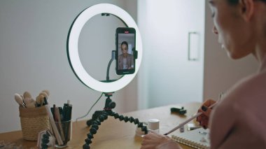 Vlogger kızı akıllı telefonuyla makyaj yapıyor. Yakın plan çekimde. Güzellik blogcusu cep telefonu kamerasına günlük yüz tanıma programı çekerken dudak boyuyor. Kozmetik ürünlerini inceleyen nüfuzlu kadın.