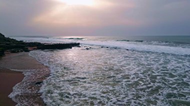 Sakin okyanus kasvetli manzara uzaklaşır. Volkanik karanlık dağ arka planına sahip derin deniz suyu. Sakin ve güzel denizin üzerindeki bulutlu gri gökyüzü. Dalgalar kumlu sahile süzülüyor Süper yavaş çekim