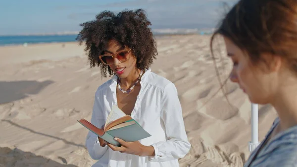 Focused girl reading book at ocean picnic. Two friends resting on sandy beach in summer sunlight. Attractive african american studying enjoying sea breeze. Love partners spending time relax on coast.