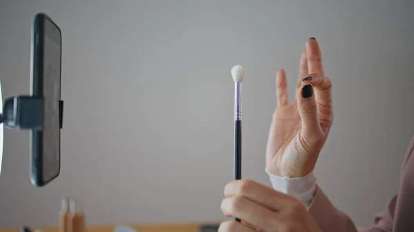 Visagiste fingers cosmetics brushes review making at house closeup. Unknown blogger showing brush gesturing to smartphone camera indoors. Woman influencer hands reviewing visage equipment to cellphone