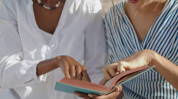 Closeup hands reading book on summer beach. Cheerful women enjoying ocean shore vacation discussing pointing page. Smiling multiethnic couple spending weekend together. Friendship bonding concept.
