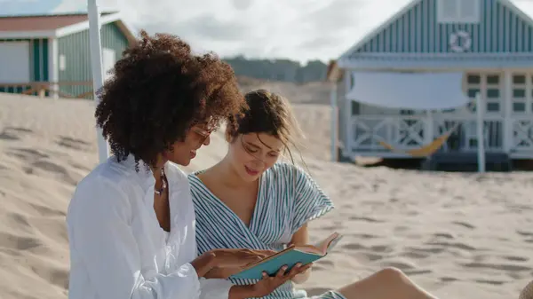 Two girls reading book on summer beach picnic. Happy lesbian couple talking enjoying novel in sunlight. Carefree multiethnic friends discussing literature on sandy coast. Vacation weekend concept.