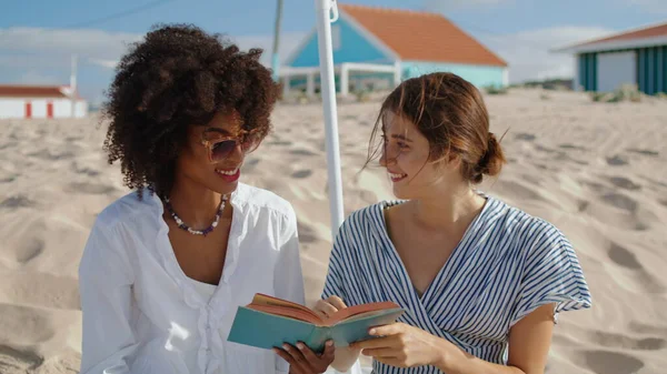 Friends enjoying picnic book at sandy shore. Cheerful girls having fun reading novel in summer sunlight. Beautiful multiethnic partners rest together on beach. Relaxed lgbt couple date at ocean coast